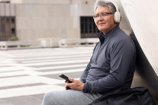 Senior man sitting on ledge against curved sculpture in city plaza holding smartphone, copy space