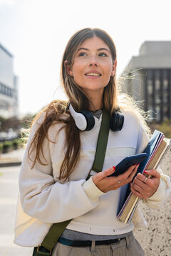 Female student standing on city walkway carrying notebooks and smartphone with headphones on neck