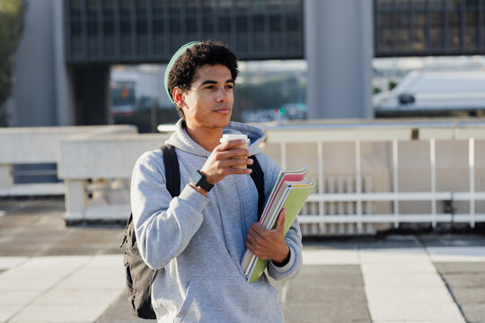 Male student standing on rooftop near railing holding coffee cup and notebooks wearing backpack