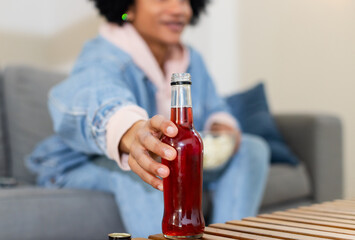 African American woman wearing denim jacket sitting on couch holding red drink bottle and popcorn