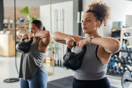 Diverse female friends performing upright row with kettlebells in fitness studio near dumbbell rack