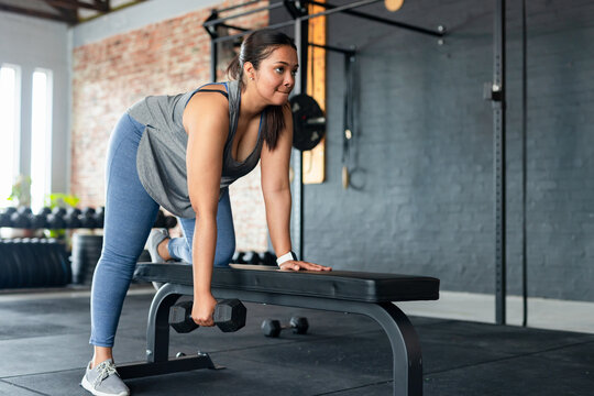 Woman in grey tank top, blue leggings lifting dumbbell on bench near dumbbell rack in gym
