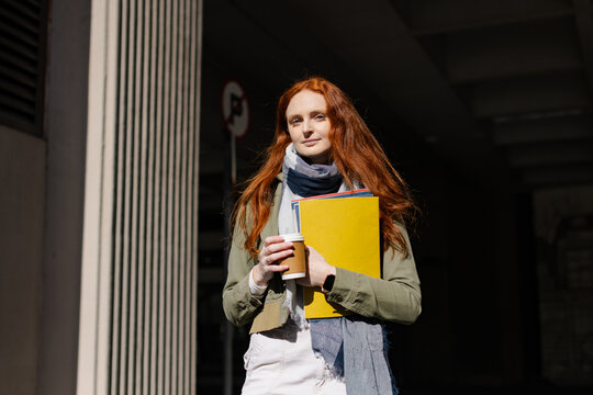 Redheaded woman carrying coffee yellow folder papers red notebook under walkway wearing gray scarf
