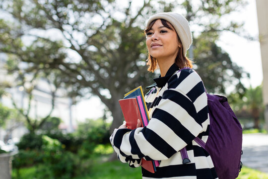 Female student standing on campus path holding textbooks wearing knit beanie, backpack, copy space
