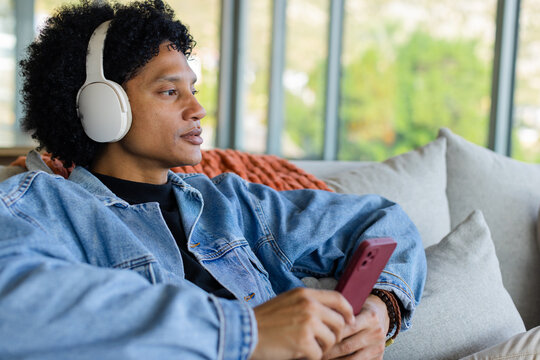 African American man reclining on sofa by window at home holding smartphone wearing headphones - Powered by Adobe