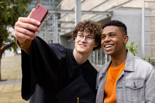 Diverse male friends wearing gown taking selfie in campus yard with mortarboard and red smartphone