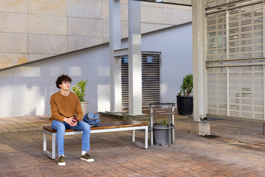 Man sitting on bench wearing sweater using smartphone with duffel bag at courtyard, copy space