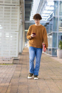 Man walking along covered campus corridor holding smartphone and red folders near glass facade