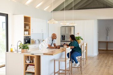 Diverse couple checking smartphone while holding coffee mugs at wooden countertop island in kitchen