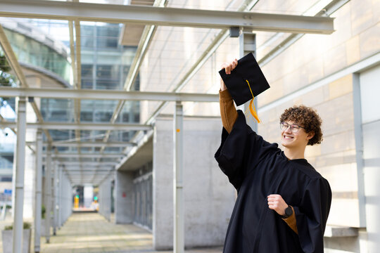 White male graduate standing under canopy in black gown holding cap overhead smiling, copy space