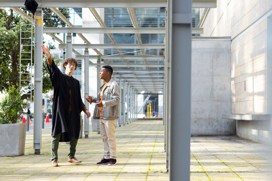 Diverse male friends tossing mortarboard cap under metal walkway with planter and traffic cones