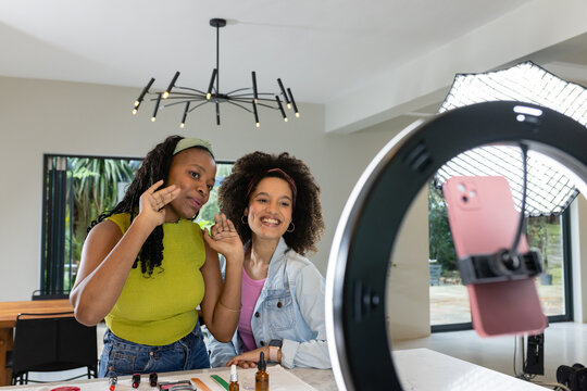 Diverse female friends standing at dining table filming makeup tutorial with ring light smartphone