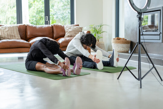 Diverse couple performing forward bend on green mats in living room beside potted plant, ring light - Powered by Adobe
