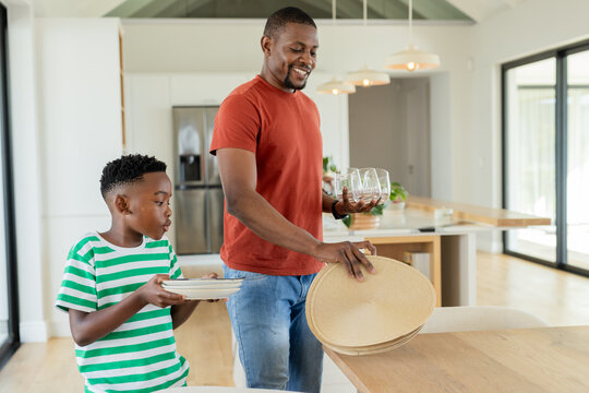 African American father and son placing straw placemats, white plates and wine glasses in kitchen