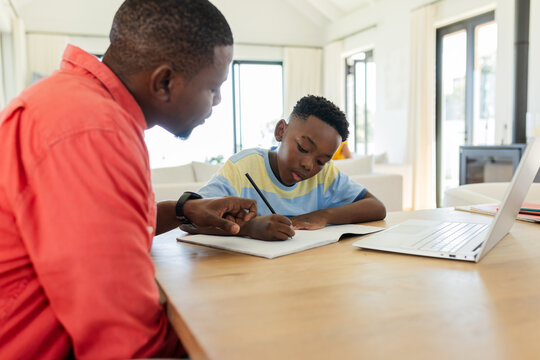 African American father and son sitting at wooden table at home, writing in notebook beside laptop