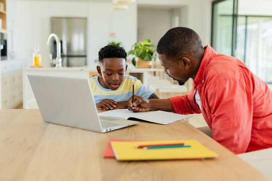 African American father and son studying homework at kitchen table using laptop writing in notebook