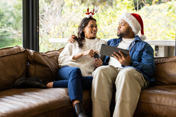 Diverse couple sitting on sofa in living room, wearing festive headbands, holding tablet and remote