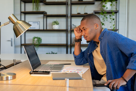 African American man resting hand on forehead while analyzing charts on laptop at desk, copy space