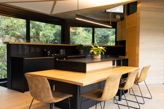 Wooden island doubling as dining table with leather chairs, potted plants and tall windows