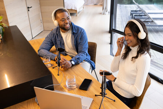Diverse hosts speaking into tabletop mics at home studio on wooden table with laptop, headphones