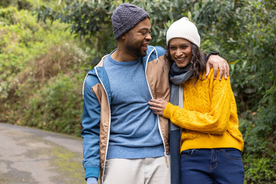 Diverse couple strolling arm-in-arm on trail wearing beanies, yellow sweater, jacket, gray scarf