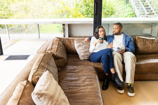 Diverse couple sitting on leather sectional sofa in living room holding coffee mugs, copy space
