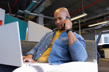 African American man talking on smartphone while using laptop on office sofa wearing smartwatch