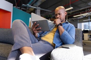 African American man reclining on sofa in office holding tablet supporting chin near smartphone