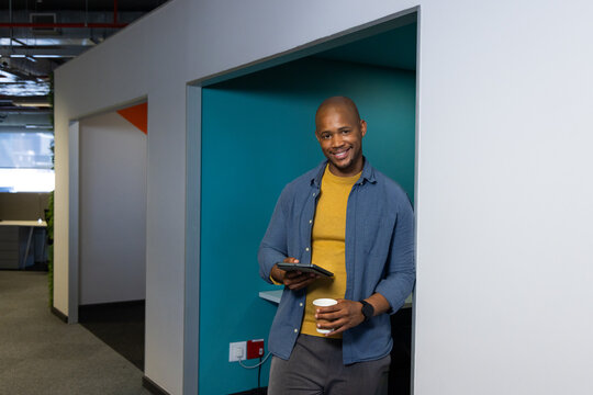 African American man leaning in office alcove holding tablet and coffee cup beside power outlet