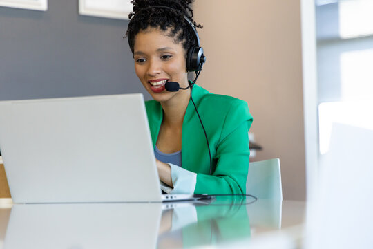 African American woman typing on silver laptop at glass table in office wearing headset in meeting