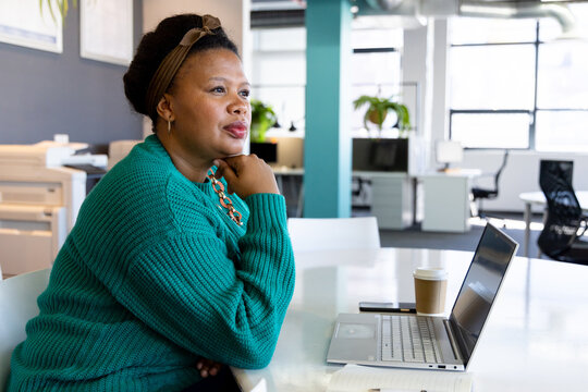 African American woman in teal sweater working on laptop while grabbing coffee in open-plan office
