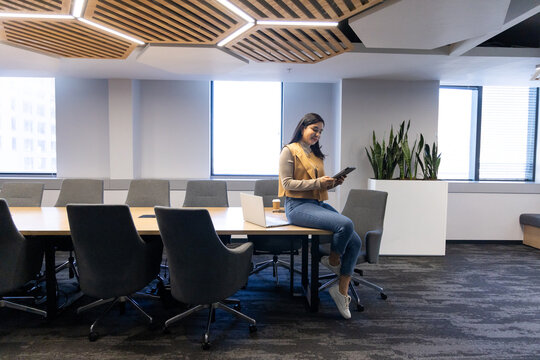 Woman holding tablet while sitting on meeting room table edge with laptop, copy space