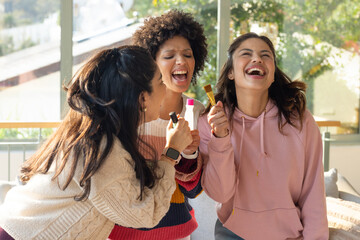 Diverse female friends laughing and singing with makeup brush bottle lipstick on sunlit sofa