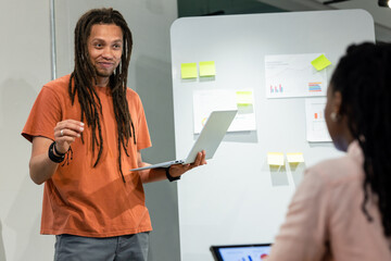 Team leading, gesturing in office wearing casual attire at board with charts, sticky notes, laptops