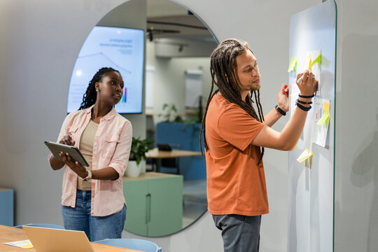 African American team placing sticky notes and checking tablet on whiteboard in office, copy space