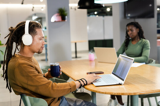 African American coworkers typing on laptop at table in coworking space wearing headset holding mug - Powered by Adobe