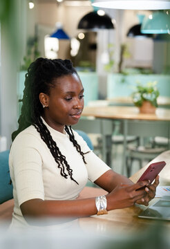 African woman sitting in coworking lounge wearing white top holding rose smartphone near laptop