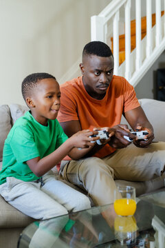 African American father and child boy sitting on sofa playing games with gamepads and orange juice