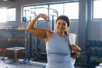 Female flexing right biceps, posing and smiling in gym with towel, light blue top, kettlebells