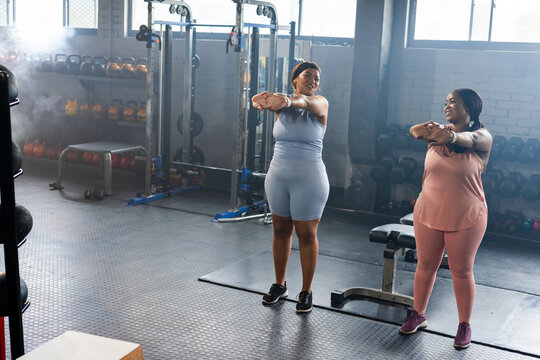 Diverse female friends stretching arms across chest in matching activewear at gym with kettlebells