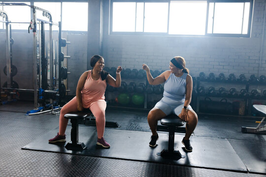 Diverse female friends sitting on benches in gym, fist bumping by dumbbell rack, kettlebells nearby