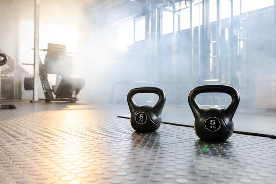 Two black kettlebells are resting on textured rubber floor in commercial gym, with sunlight haze