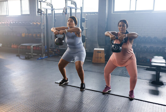 Diverse female friends wearing matching workout sets performing kettlebell goblet squats at gym