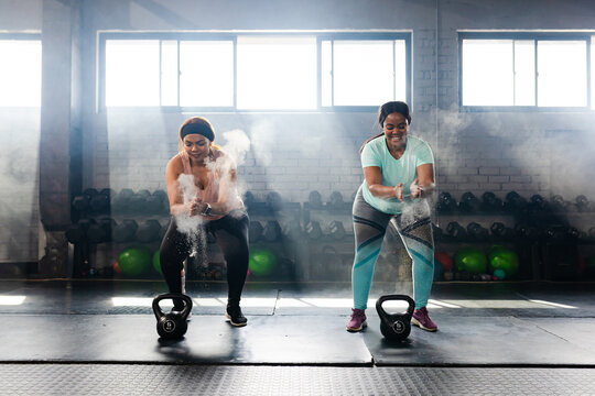 Diverse female duo in athletic wear clapping and rubbing chalk into hands by kettlebells in gym