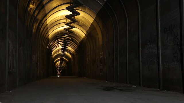 Perspective view inside a dark, long pedestrian tunnel with yellow overhead lights. The gritty urban underpass features arched walls covered in graffiti and a mysterious, eerie atmosphere.


