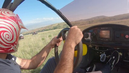 Man with red helmet drives yellow buggy on grassy hills. Action shot features driver steering and close-up view of rugged tire rolling over dry grass terrain.

