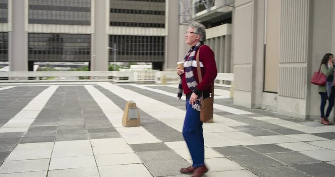 Senior man entering plaza carrying coffee, sipping while walking as senior woman on phone following