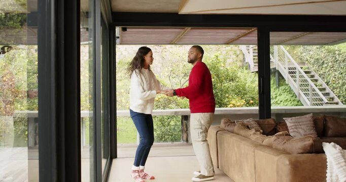 Diverse couple clasping hands and dancing with rhythmic steps beside glass wall near suede couch