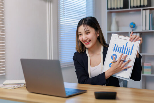 Smiling young businesswoman presenting business growth charts on a clipboard during a remote online video call meeting using a laptop - Powered by Adobe