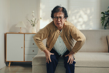 Senior woman sitting on a sofa, experiencing sharp knee pain and discomfort. She is holding her...